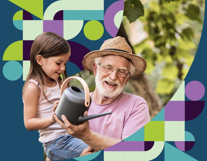 White old grandfather with white beard and a gardening hat holding his grand daughter while watering a plant
